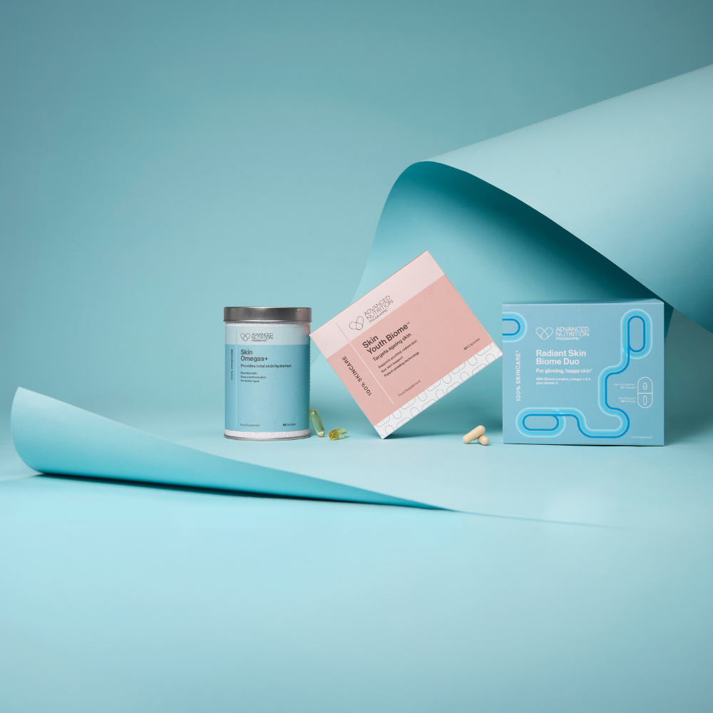 A clean studio product shot showing a light blue supplement canister, a pink and white product card, and a matching light blue box on a pale blue background with curled paper props. The brand name visible on the packaging appears to be 'Gourmet Health'.