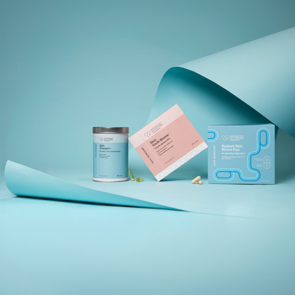 A clean studio product shot showing a light blue supplement canister, a pink and white product card, and a matching light blue box on a pale blue background with curled paper props. The brand name visible on the packaging appears to be 'Gourmet Health'.
