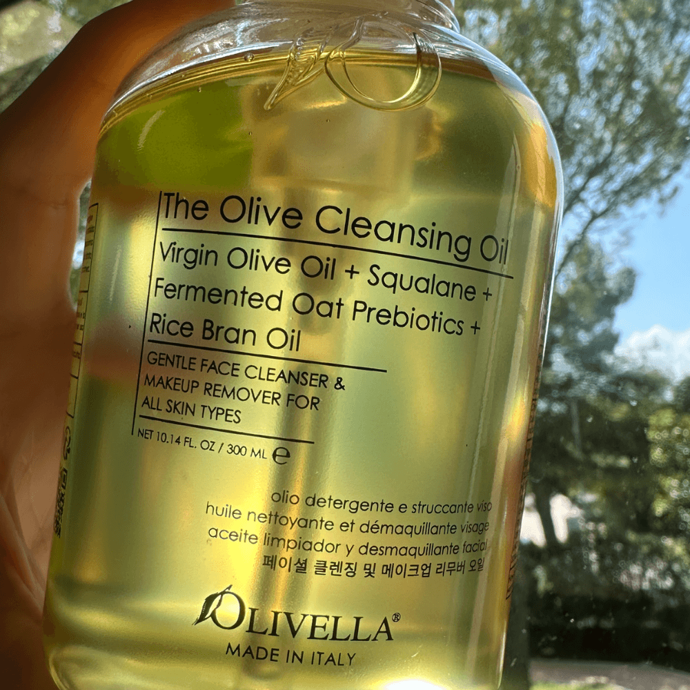 A hand holding a clear plastic bottle of Olivella The Olive Cleansing Oil against a bright outdoor background with trees and sky visible through the transparent container.