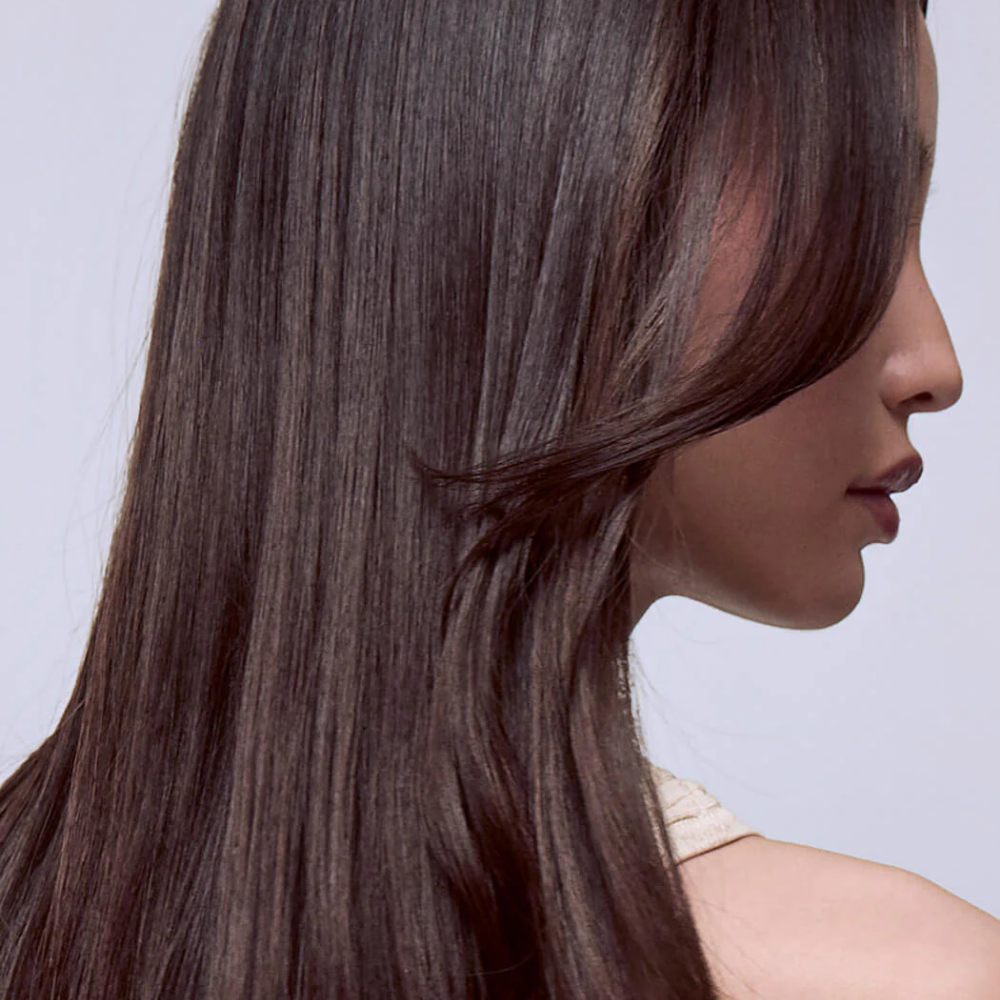 A close-up side profile of a woman with long, straight, dark brown hair, showing the texture and shine of the hair against a light gray background.