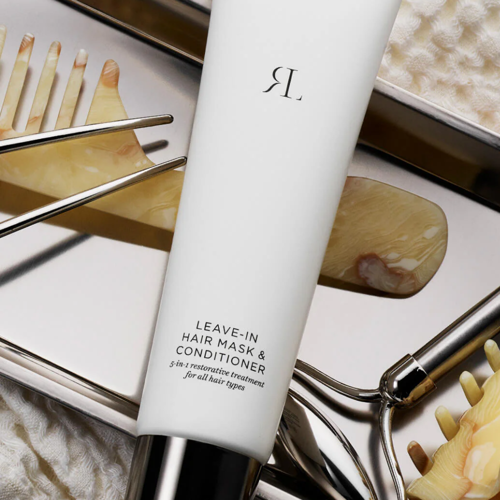 A white tube of hair mask and conditioner with a metallic silver cap, placed diagonally among hair care tools and accessories on a light surface. The tube is labeled 'R.
