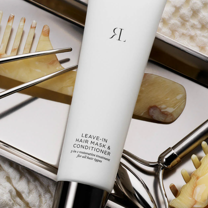 A white tube of hair mask and conditioner with a metallic silver cap, placed diagonally among hair care tools and accessories on a light surface. The tube is labeled 'R.