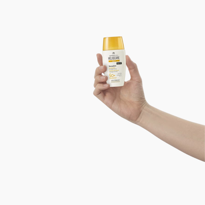 A hand holding a bottle of HelioCare sunscreen against a plain white background.